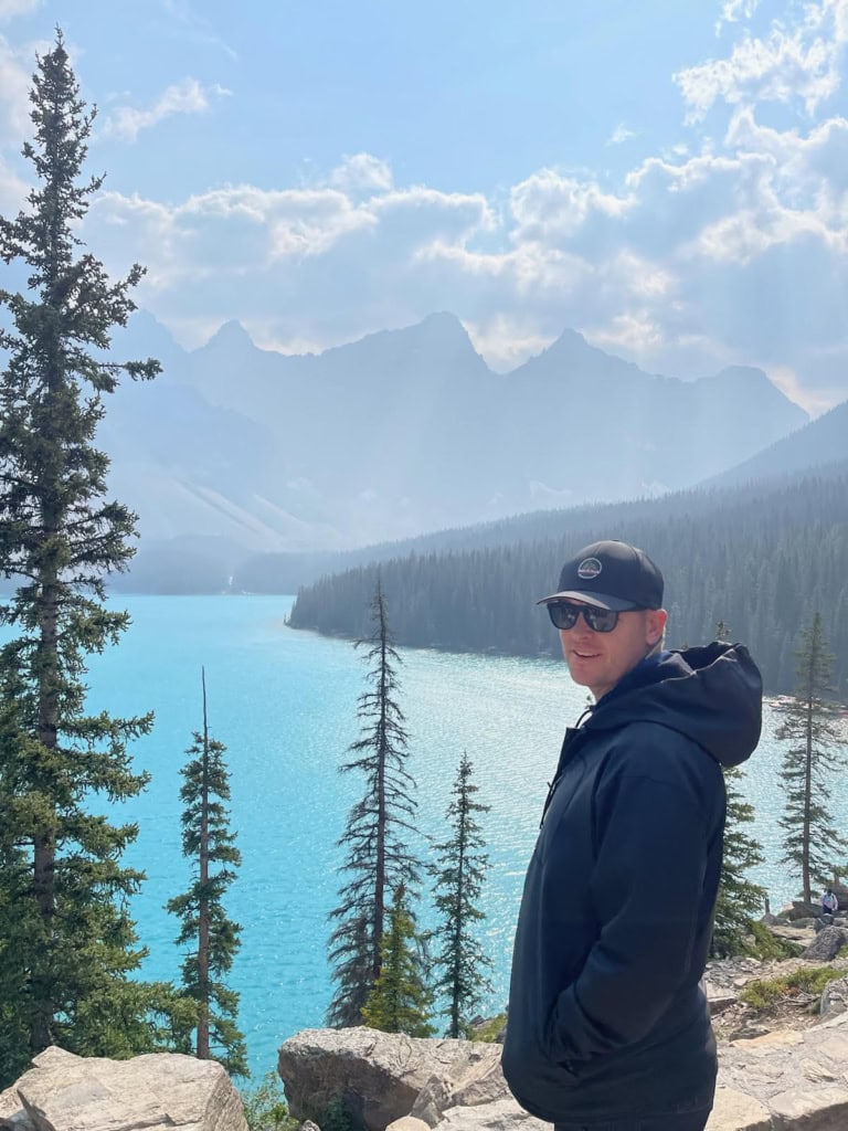 A man wearing a hat and jacket stands in front of mountains and a lake