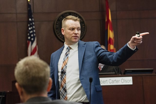 A man wearing a suit and tie stands in a courtroom as he presents a case to a jury